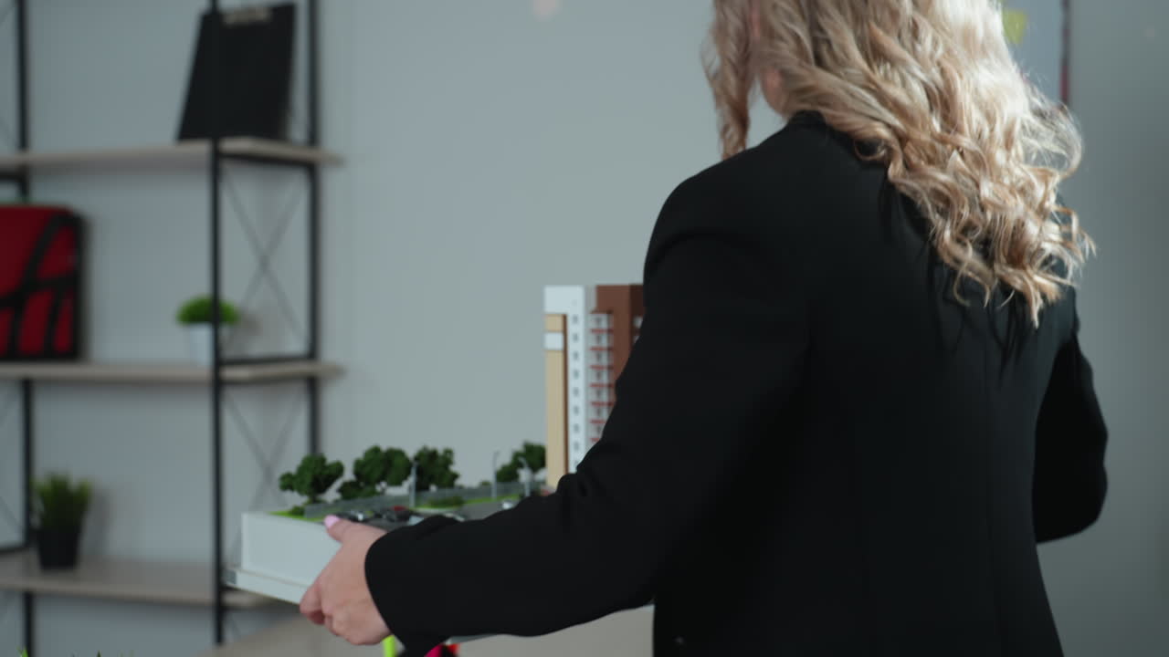 Close up of focused real estate agent holding architectural building model and carefully placing it on desk in modern office environment with shelves and clean background