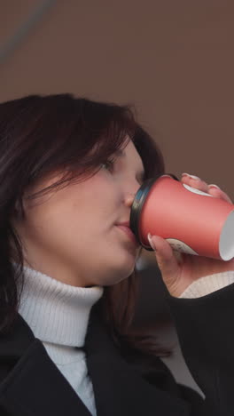 woman in black coat, sitting under patio, takes a sip of her tea cup, bursts into laughter, and gently places the cup back on table, a partial laptop is in front of her