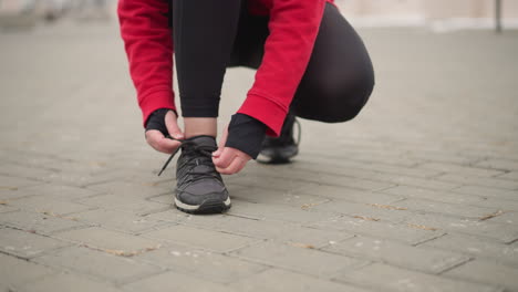 athlete tying her right shoelace while squatting on interlocked pathway wearing red hoodie, gloves, and black leggings, focused preparation on outdoor fitness routine