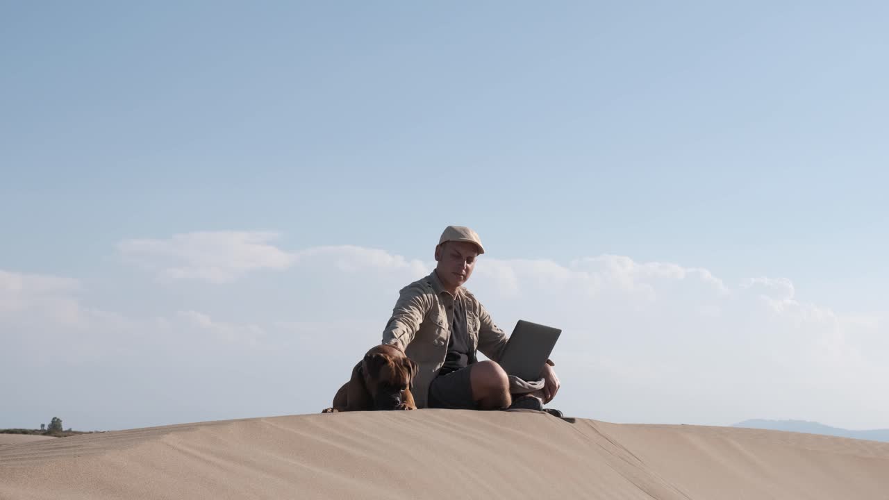 Male traveler sitting on sandy dune with dog and using laptop during vacation in sunny day