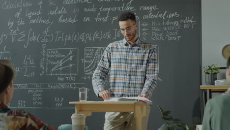 a young man lecturing students in a classroom with a blackboard full of scientific equations
