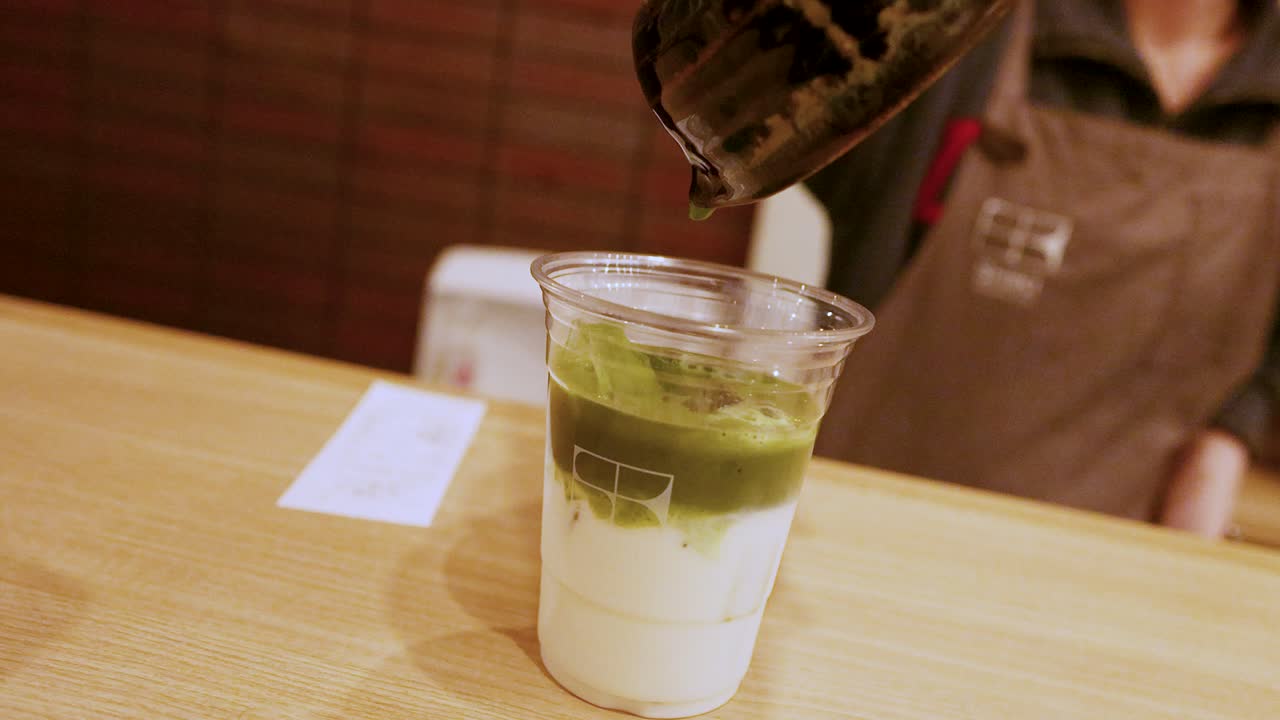 Um barista despeja chá matcha verde vibrante em um copo plástico com leite e gelo