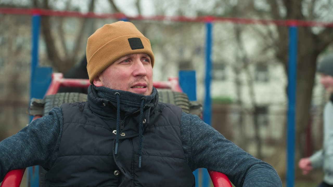 A focused coach sits on workout equipment, looking intent, in the background, two boys are seen playing football happily