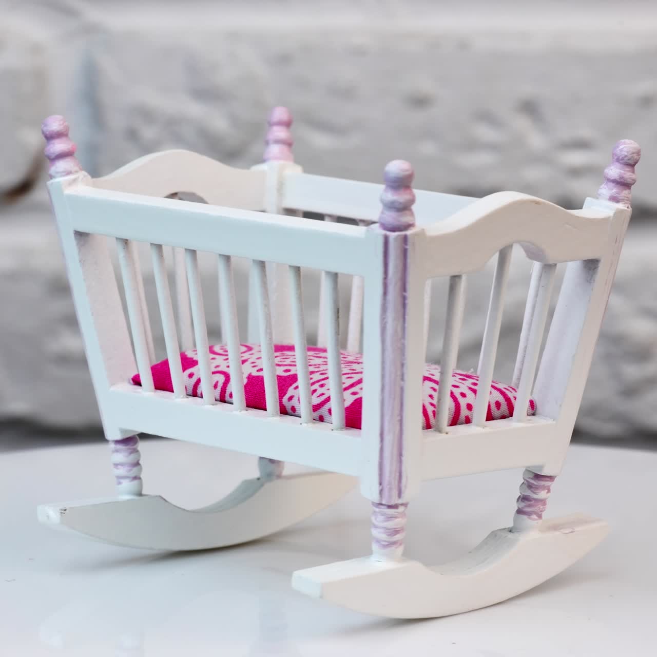 Beautiful toy cradle on a turning table. White baby crib made of wood with pink mattress as a piece of toy furniture. White brick wall at the backdrop