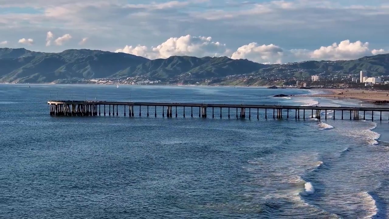 Ripresa aerea cinematografica con drone 70mm del Molo di Santa Monica, che ruota lentamente sull'Oceano Pacifico, con la costa di Los Angeles e le montagne di Santa Monica in lontananza.