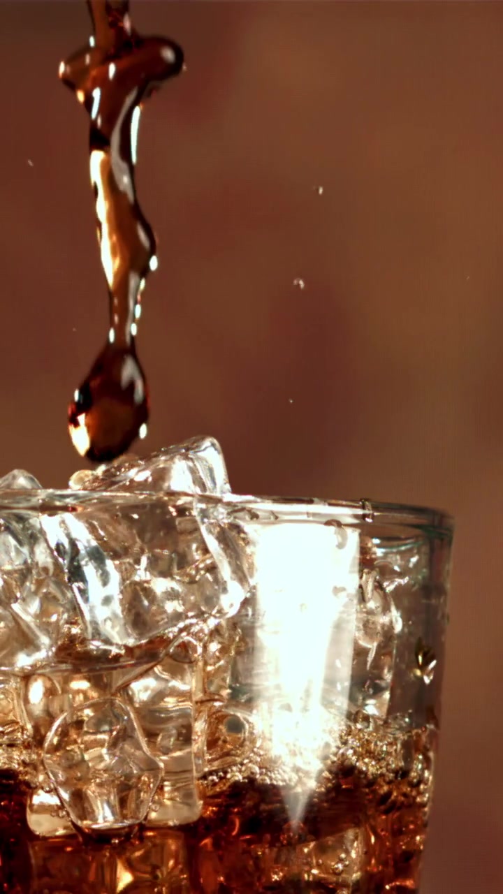 Versamento al rallentatore di liquido scuro in un bicchiere con cubetti di ghiaccio