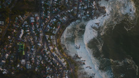 Aerial-birds-eye-overhead-top-down-view-of-big-waves-rolling-on-sea-coast.-Buildings-in-residential-borough-along-shore.-Cape-Town,-South-Africa