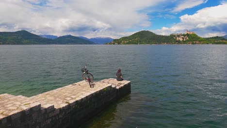 unrecognizable solitary worried woman with bicycle behind sitting on jetty edge of maggiore lake, italy