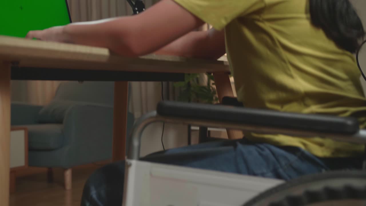 Back View Of Asian Kid Girl Sitting In A Wheelchair Stretching While Using Laptop Computer With Mock Up Green Screen At Home