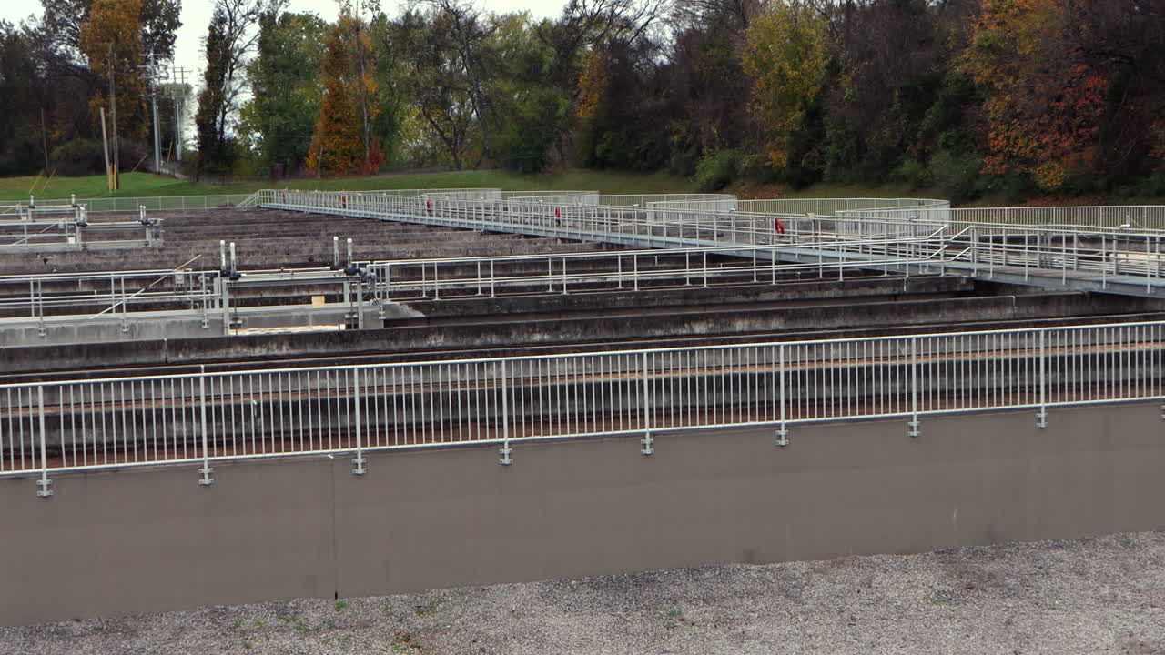 Elevated side view of flowing waste water moving through aeration basins with metal guardrails and a metal crosswalk spanning the length of the aeration basins. Trees line the edge of the basins