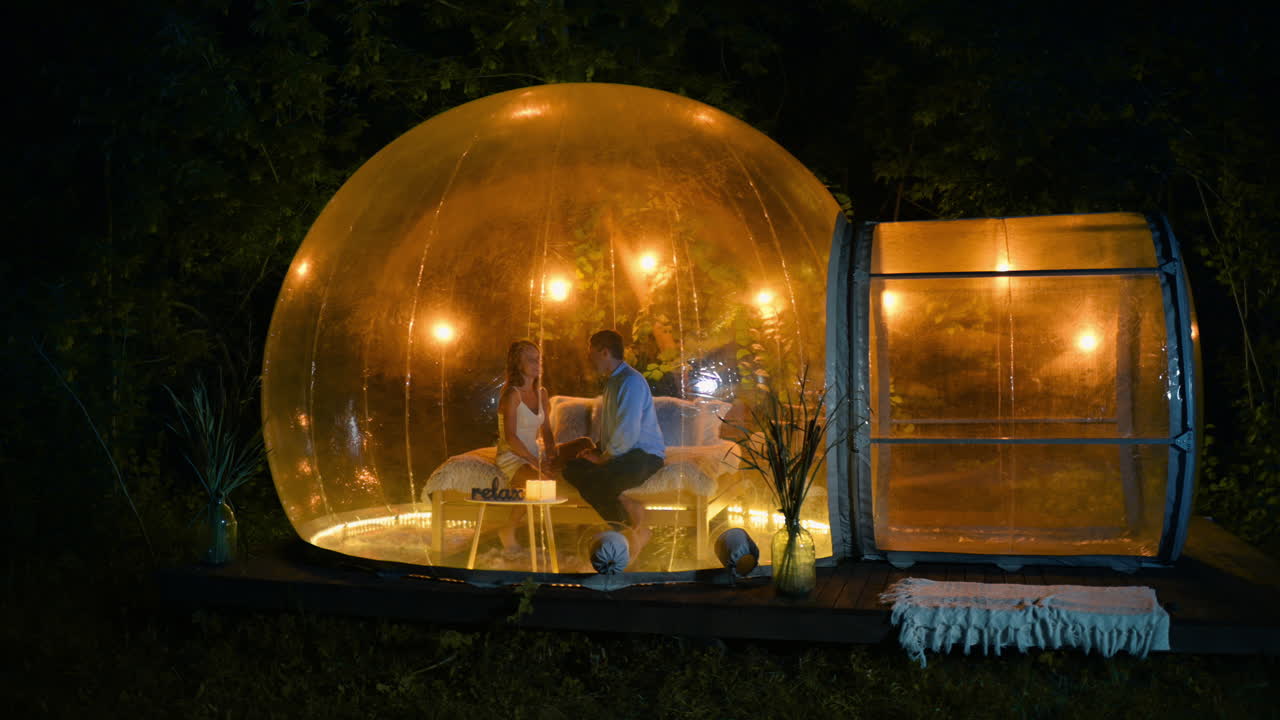 Et par, der snakker, mens de sidder i et gennemsigtigt boble-telt med belysning på glamping om natten. Frodig skov i baggrunden.