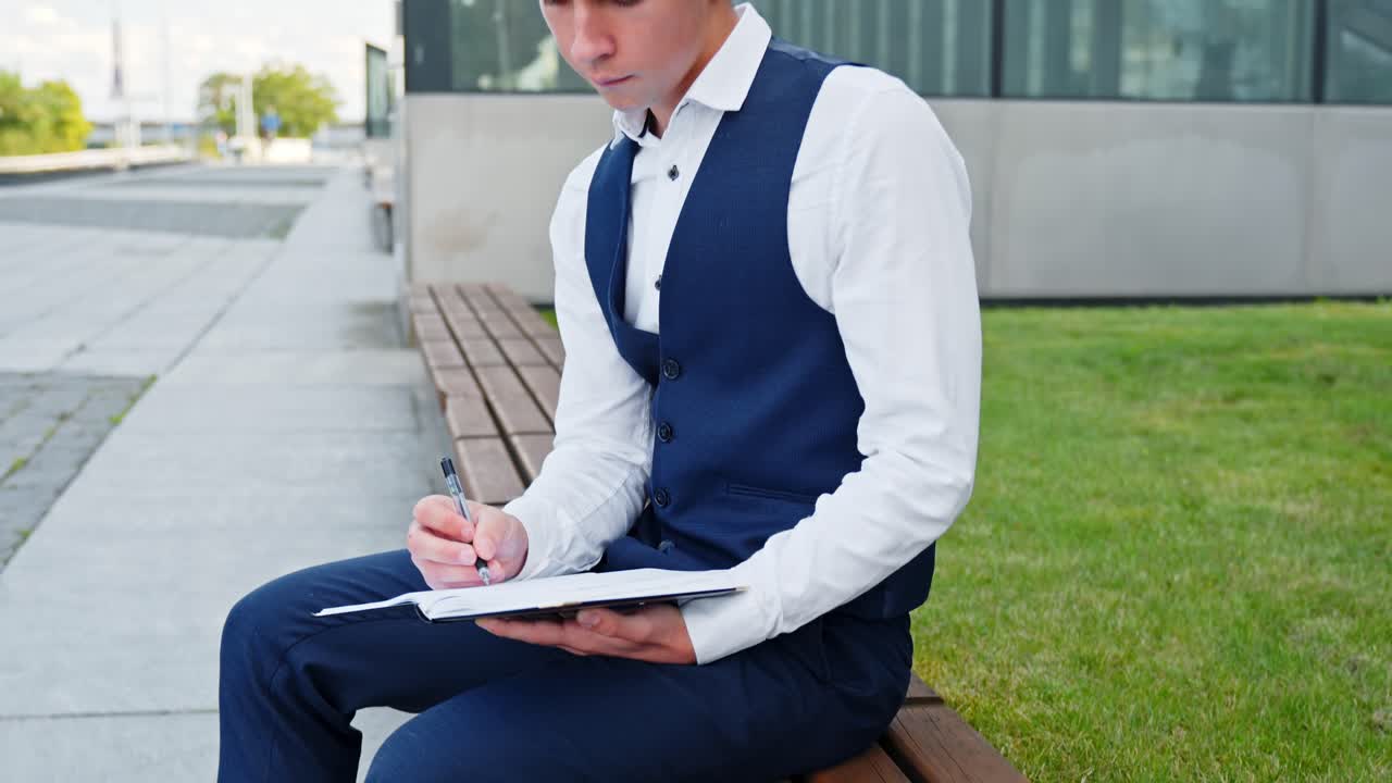 Man in suit writing on notepad outdoors, serious focus and intent noted