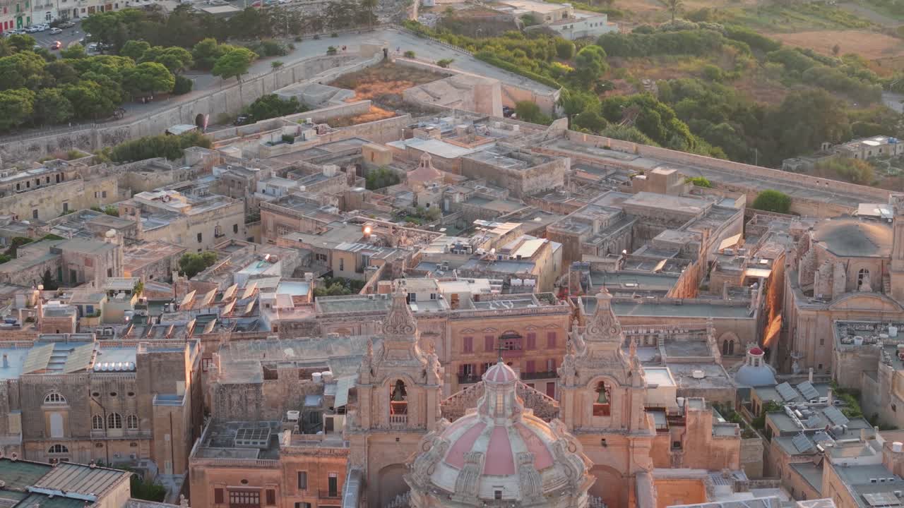 A close aerial view of St. Paul's Cathedral in Mdina, Malta, highlighting its baroque dome and twin bell towers. The warm evening light emphasizes the intricate stonework