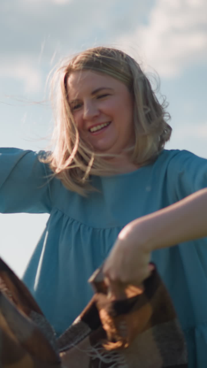 A woman smiles slightly as she holds a plaid scarf, with the wind gently blowing it in a serene outdoor setting. The scene captures a joyful and carefree moment under a bright sky