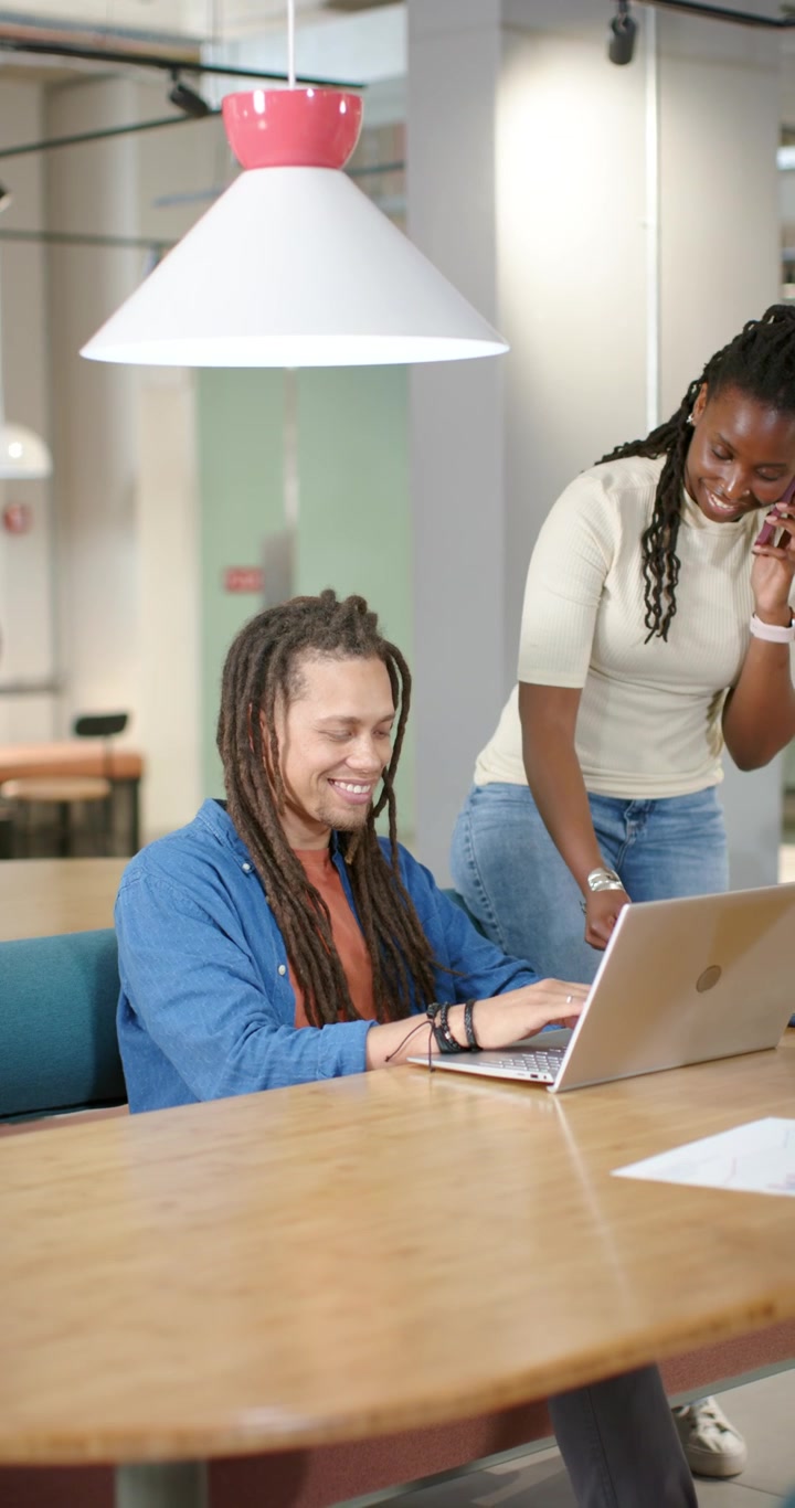 Vertical video: African coworkers at table woman coming right calling docs, man reviewing laptop
