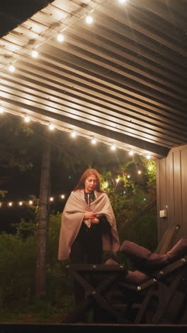 mujer se sienta en un sillón en el balcón. descanso y tranquilidad cerca de la naturaleza en una casa alquilada para visitantes. patio iluminado con lámparas brillantes rodeado de denso bosque por la noche