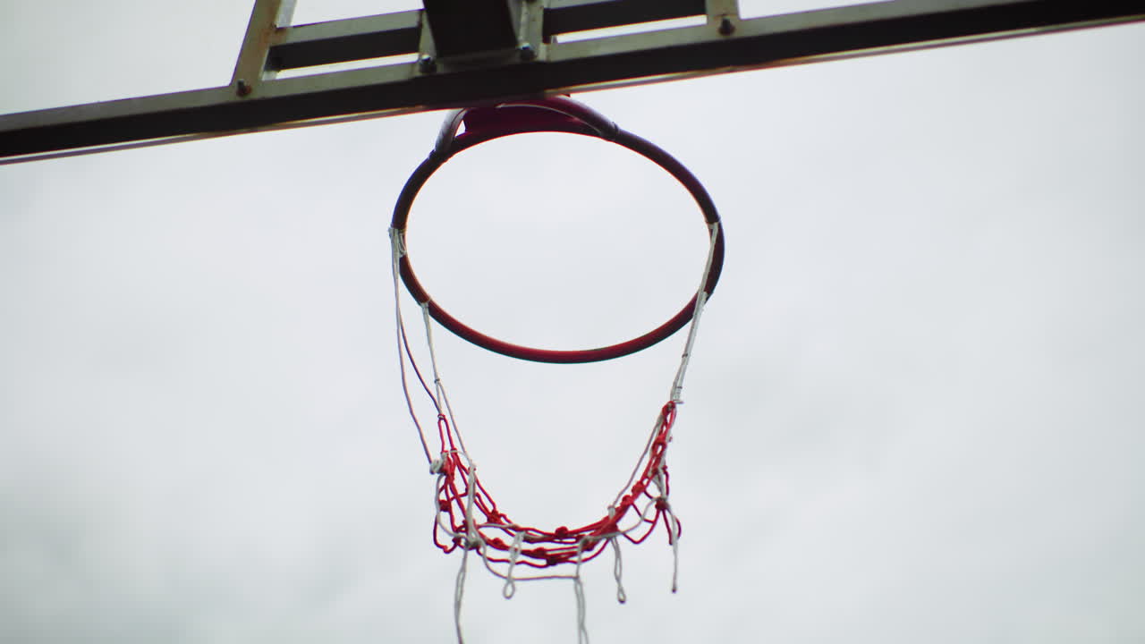 Canestro da basket con pallone sospeso a mezz'aria, rete rossa sfilacciata appesa al bordo arrugginito sotto un cielo nuvoloso, pallone che descrive un arco pulito verso il centro, un lento movimento cinematografico cattura il momento del "swish" e il leggero rimbalzo