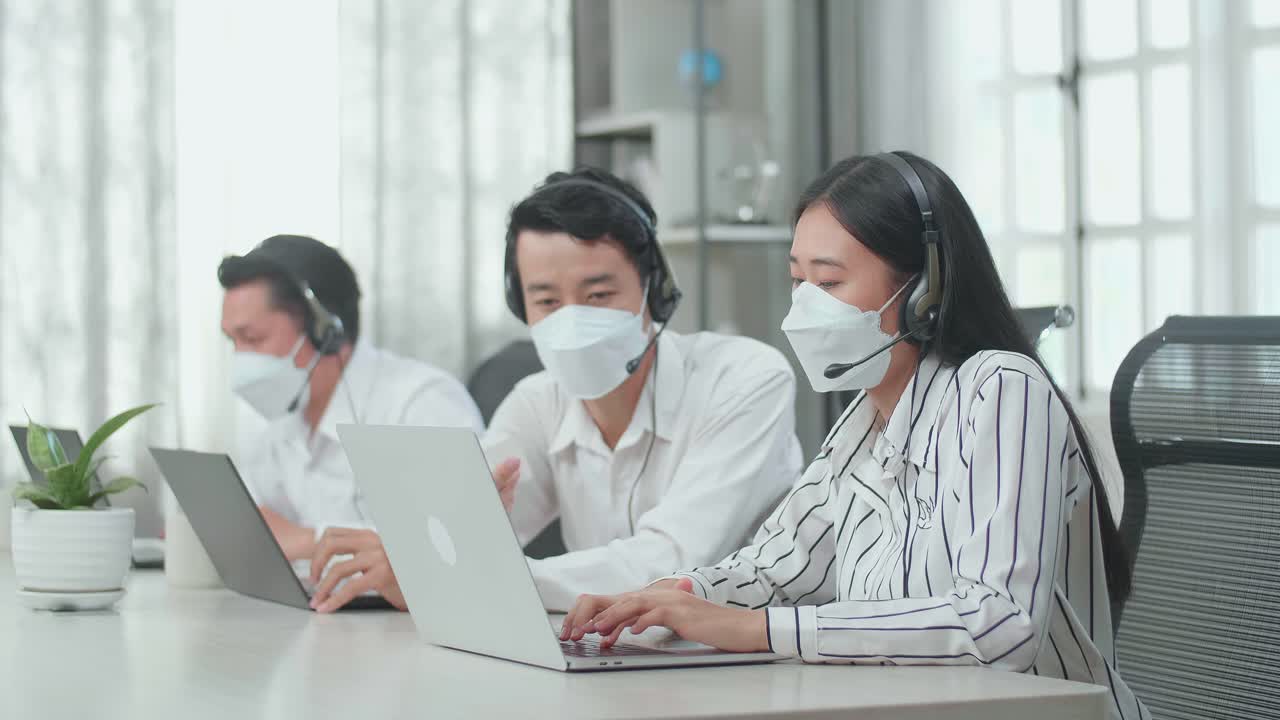 A Man And A Woman Of Three Asian Call Center Agents In Headsets And Masks Discussing Work While Their Colleague Is Speaking With Customers On The Call At The Office