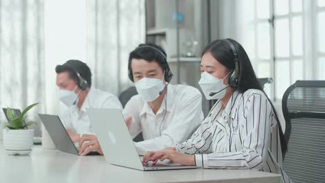 a man and a woman of three asian call center agents in headsets and masks discussing work while their colleague is speaking with customers on the call at the office