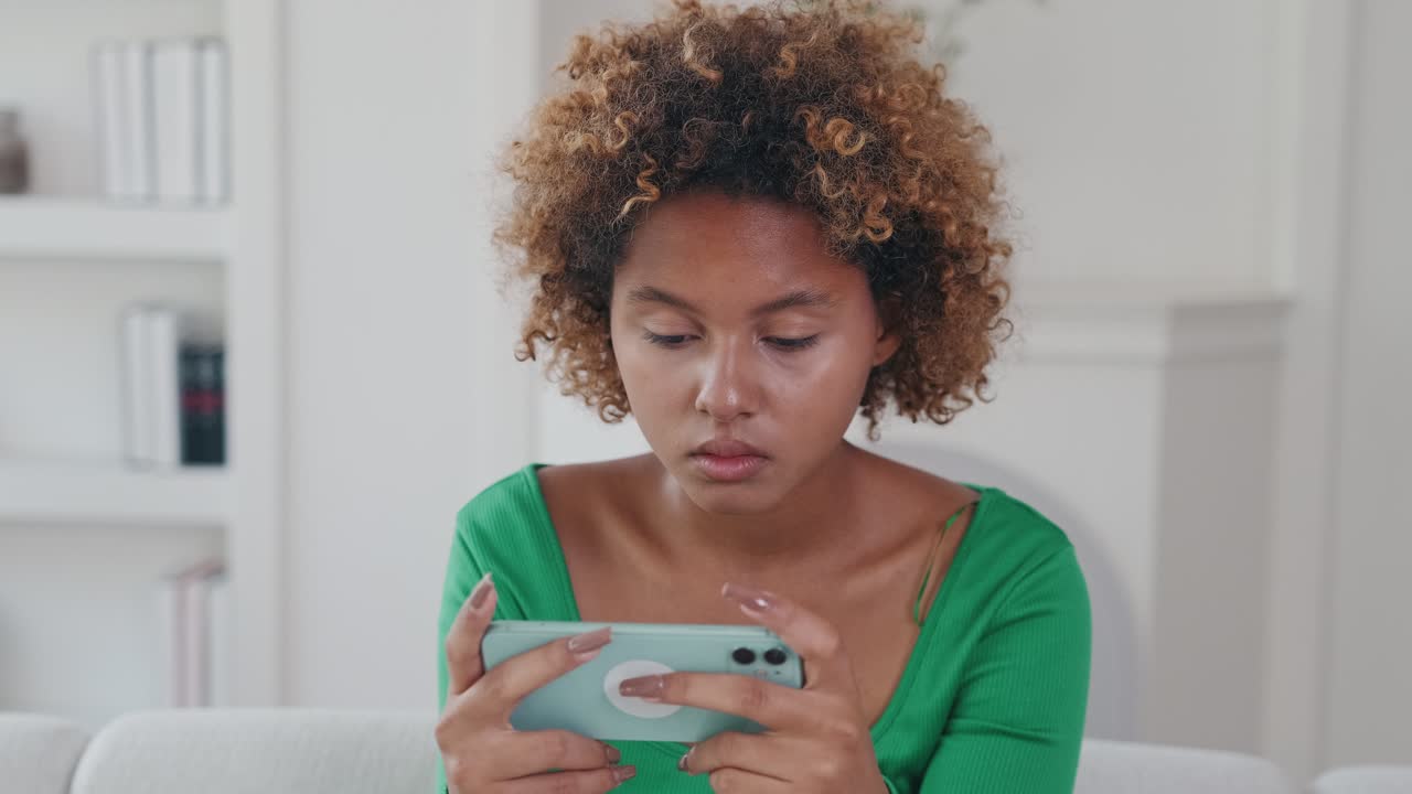 Young puzzled nervous african american woman holds phone sits on sofa