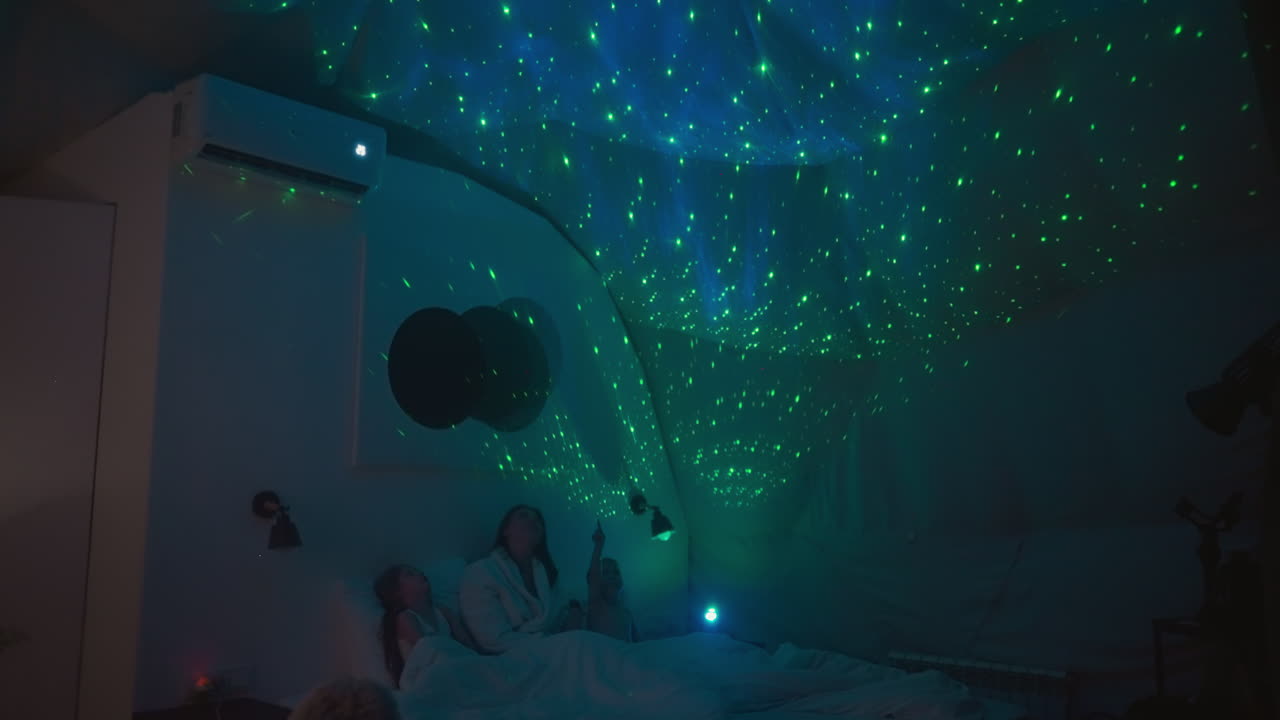 어머니와 함께 침대에 누워서 천장에 있는 하늘의 투영을 가리키며, 프로젝터에 의해 조명된 반 어두운 방, 행복한 가족을 위한 정보적인 저녁.