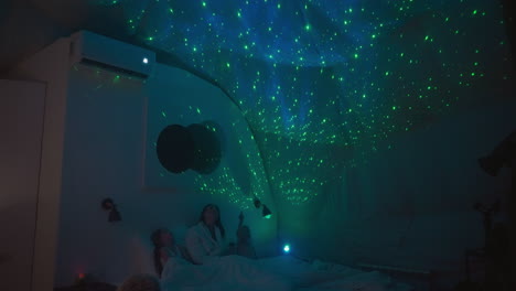 los niños con la madre se acuestan en la cama y apuntan a la proyección del cielo en el techo. habitación semi-oscura iluminada por proyector. noche informativa para una familia feliz