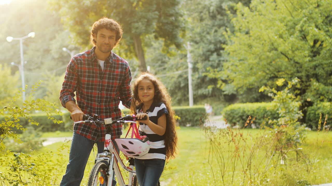 Portrait of a little girl and her father near the bike. They look at each other, then - into the camera. Smiling. Blurred background