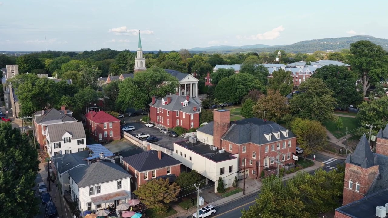 Historisk kvarter med røde murstenshuse i Charlottesville, Virginia, med et hvidt kirketårn, en søjleindgang, skifertage og en skyggefuld gade med parkerede biler. Bredt luftfoto med tilt op