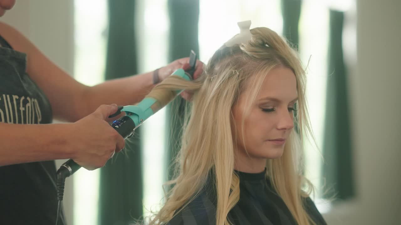 Hairdresser curls blonde woman’s hair with a hot iron, steam visible, creating locks