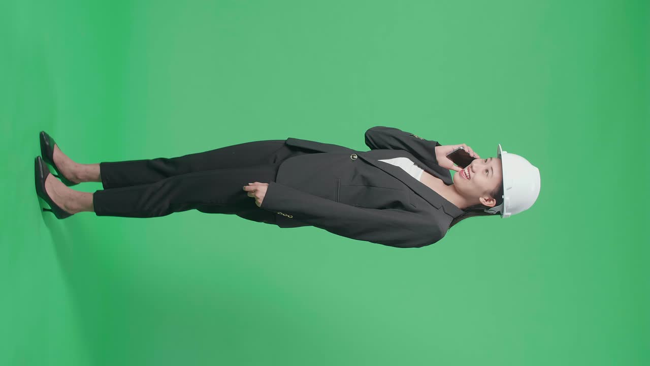 Full Body Side View Of Smiling Asian Female Engineer With Safety Helmet Talking On Smartphone In The Green Screen Studio