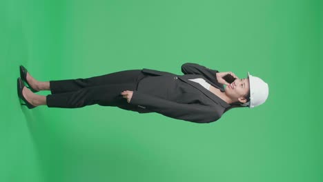 full body side view of smiling asian female engineer with safety helmet talking on smartphone in the green screen studio