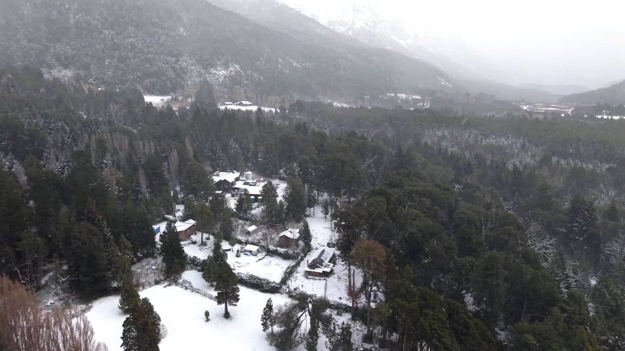 Drone captures a vast Patagonian forest blanketed by thick snowfall near Colonia Suiza, Bariloche. Snow-covered trees, cabins, and misty mountains frame the winter weather scenery from above