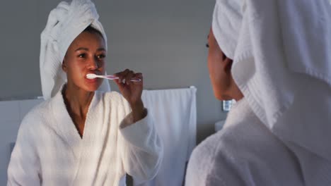 african american woman in bathrobe brushing her teeth looking in the mirror at bathroom