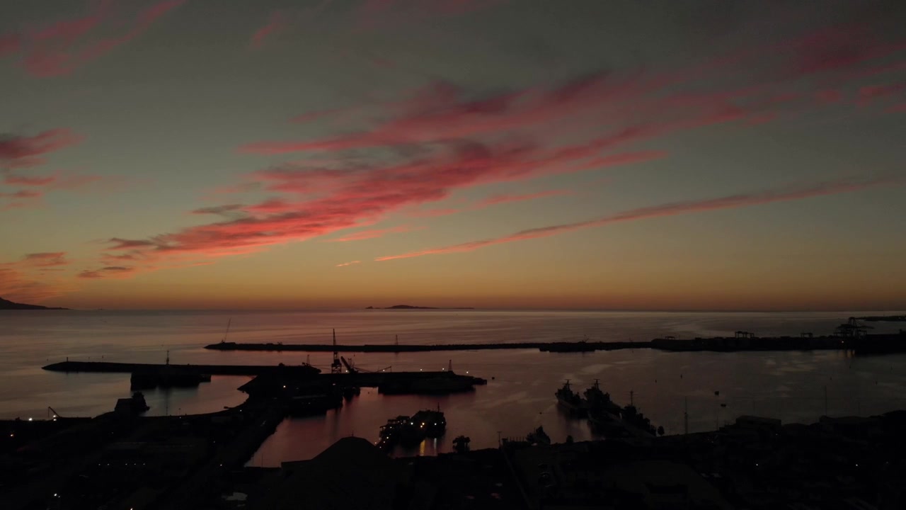 Aerial drone sunset view of harbor silhouettes calm ocean water and colorful evening clouds stretching across peaceful coastal horizon at dusk
