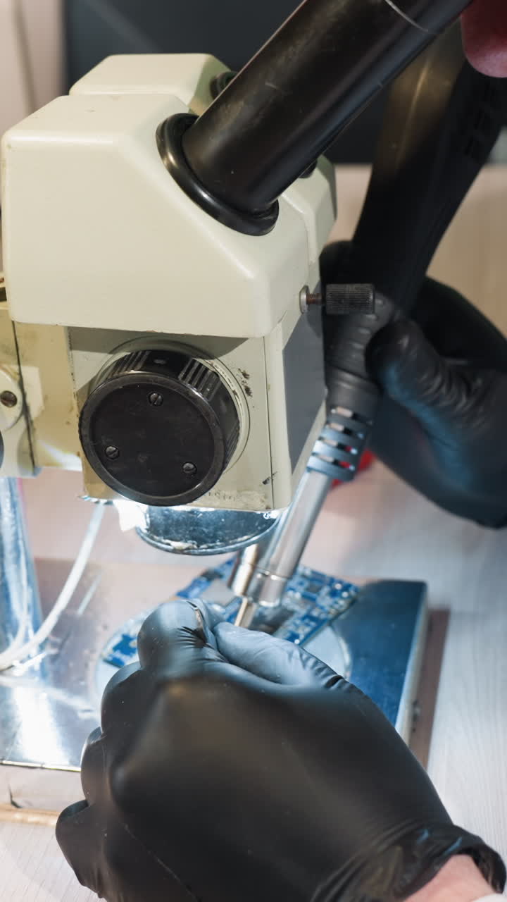 Close-up view of a technician in a white lab coat using a microscope and soldering tools to inspect and repair a circuit board