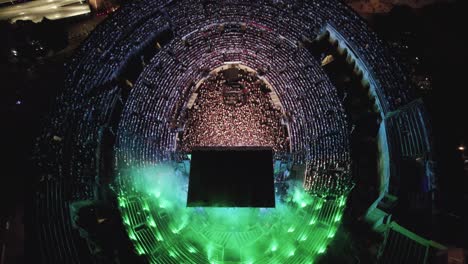 Drone-En-Las-Arenas-De-Nîmes-En-Medio-De-La-Noche,-La-Gente-Está-Viendo-El-Concierto-Y-Hay-Luces-De-Varios-Colores-Y-Ciudad