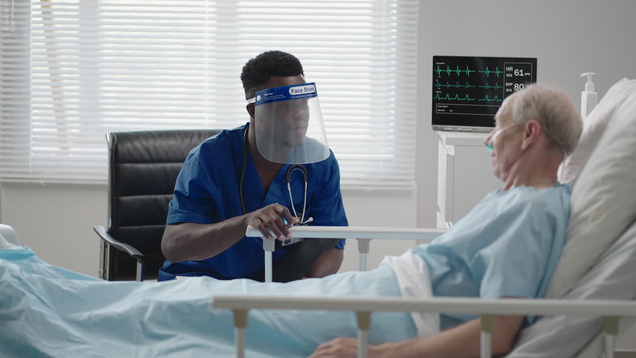 un médico negro con una máscara protectora entra y habla con pacientes con un anciano blanco acostado en una cama de hospital
