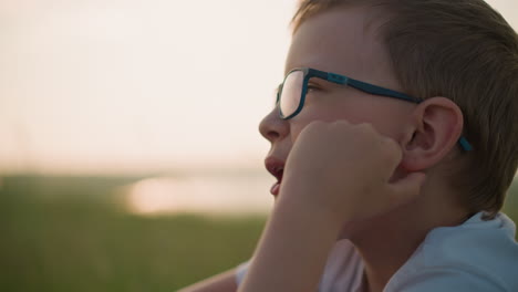close-up of a young boy wearing glasses and a white shirt, thoughtfully chewing with his hand resting on his cheek, enjoying a peaceful moment outdoors in a grassy field during sunset