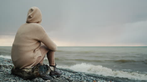 a woman in a warm sweater with a hood on her head sits on the ocean shore, where the dramatic sky and surf