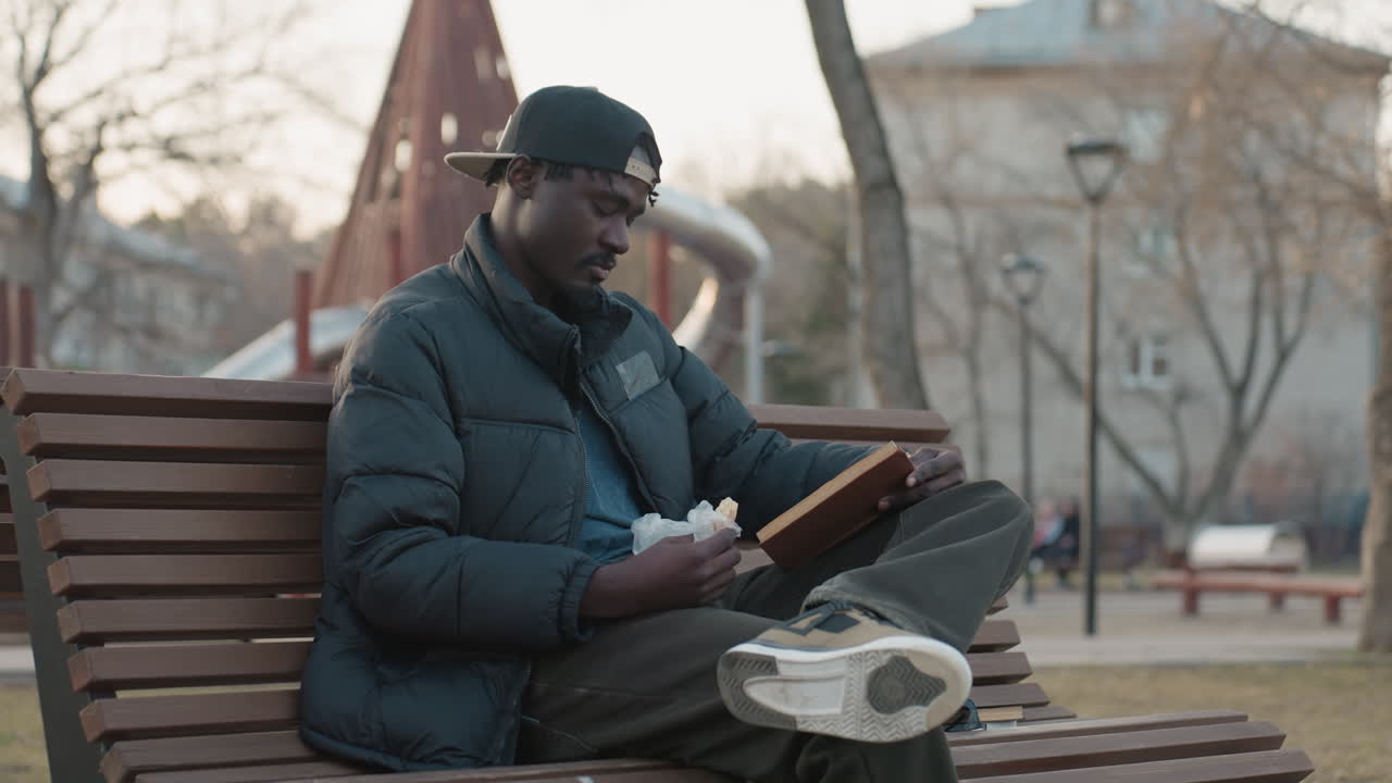 Zijaanzicht van een donkere man met een pet en pufferjack die op een bankje in het park zit en een boek leest terwijl hij een sandwich vasthoudt, tijdens een rustige middag in een stadspark omringd door kale bomen