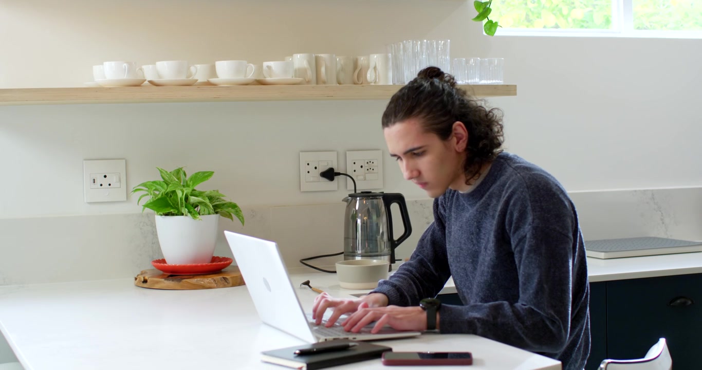 Uomo afroamericano che sposta il bollitore sulla base, lo attiva, digita sul laptop sul bancone, con spazio per il testo.