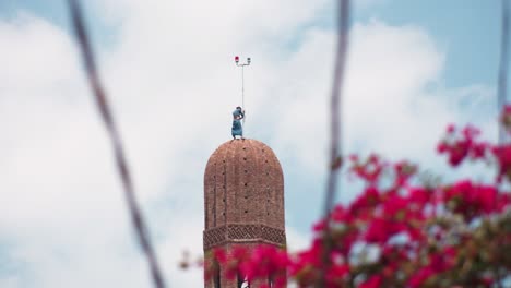 a man is trying to install copper crescent on the top of the minaret through the power cables and red flowers