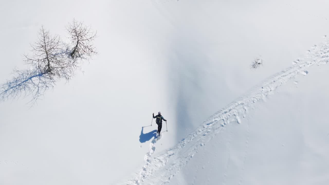 Lone hiker snowshoeing uphill on deep snow trail, leaving tracks, winter sports, solitude, endurance in pristine nature. Aerial drone top-down view, copy space