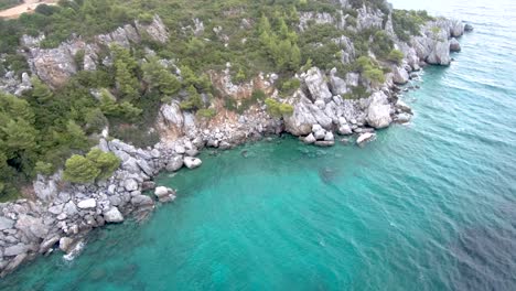 Vista-Aérea-De-Rocas-Escarpadas-En-Un-Mar-De-Cristal-Turquesa-En-La-Zona-De-Agia-Paraskevi-Halkidiki,-Grecia,-Movimiento-Circular-Por-Drones