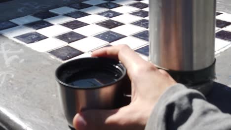 close shot of caucasian hand holding a glass of hot medicinal tea near a chess table in the sun