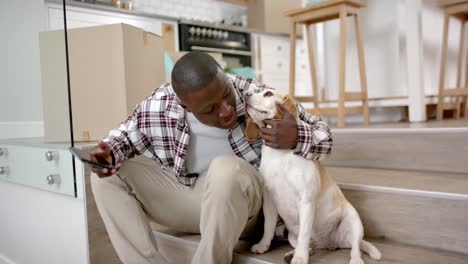 happy african american man sitting on stairs using smartphone at home, with his pet dog, slow motion