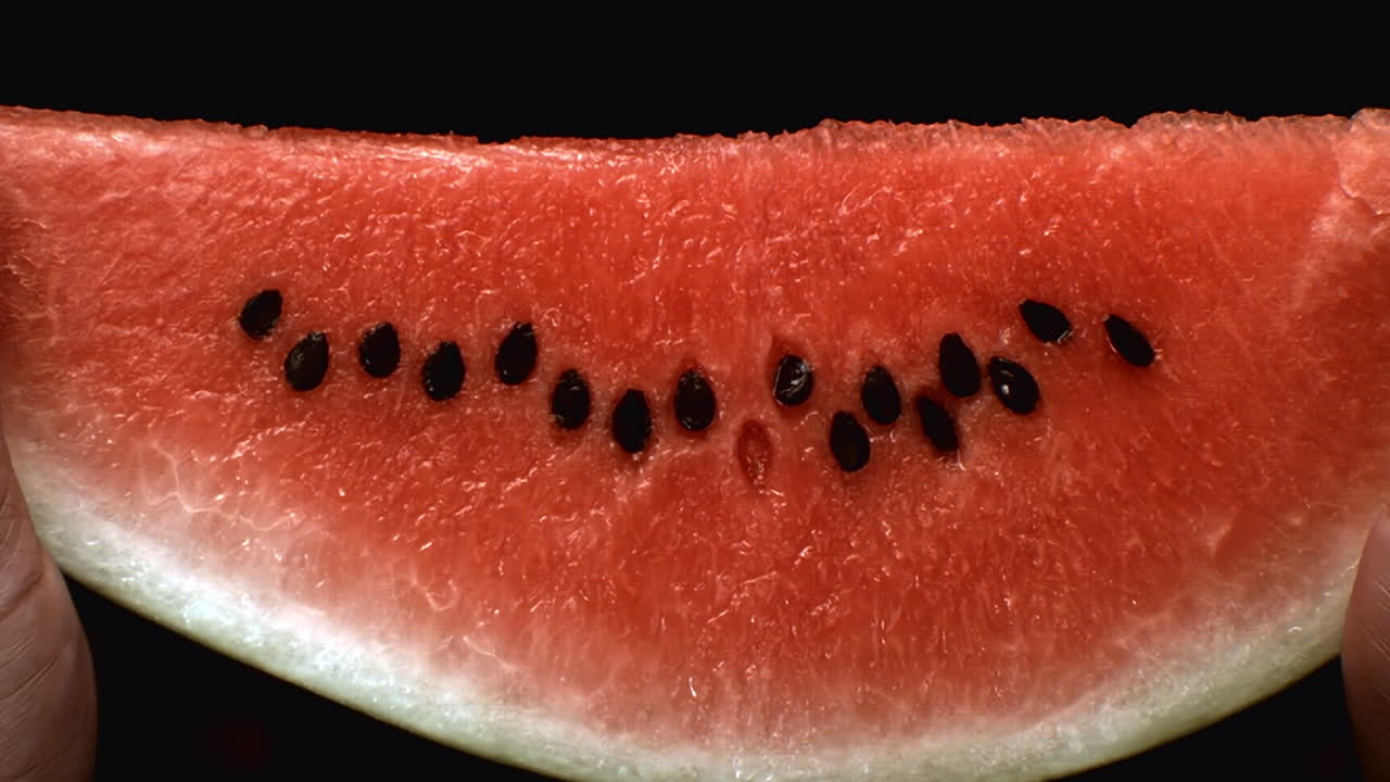 Slice of fresh watermelon with seeds on black surface showing juicy red interior, broken apart cleanly in half, water drips off in slow motion