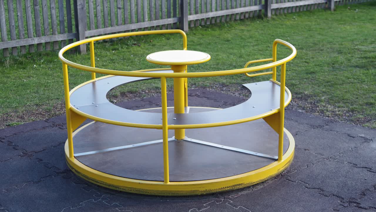 A modern yellow metal playground carousel with handrails and seats, on rubber safety flooring, with grass and a wooden fence in the background.