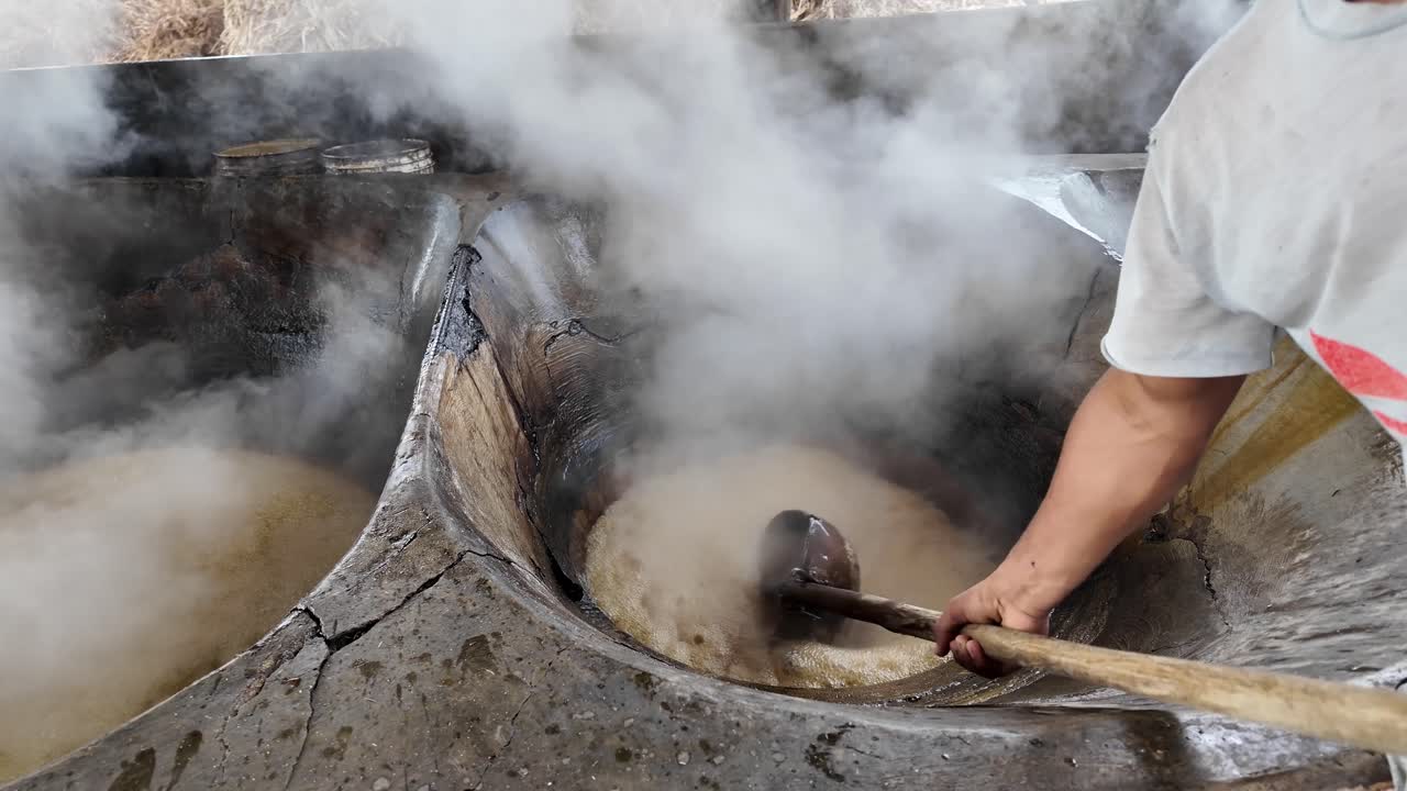 Steam rises from boiling sugar cane juice as a man scoops it from a heated vat