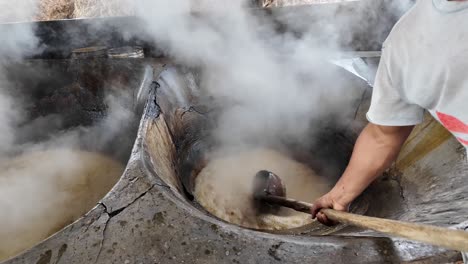 steam rises from boiling sugar cane juice as a man scoops it from a heated vat